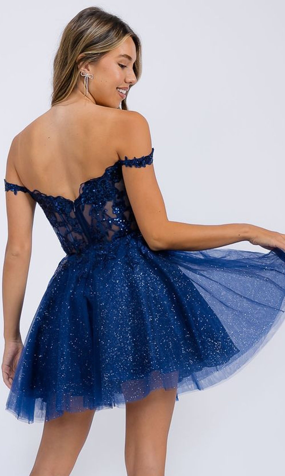 A woman models the Short Glitter-Tulle Homecoming Dress: Eureka EK138 in navy, featuring off-the-shoulder design, lace and glitter details, as she holds part of the skirt and faces slightly away from the camera.